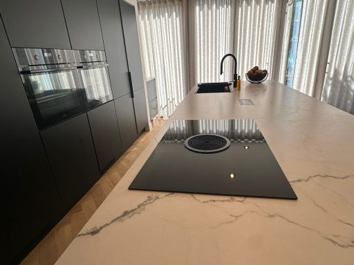 Moderne keuken met een zwart kookplaat, marmeren aanrecht en lichte gordijnen.